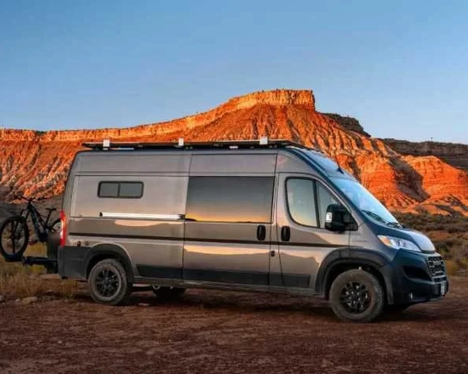 Um motorhome DeVan Ram ProMaster com um suporte para bicicletas estacionado ao pé de penhascos de rocha vermelha resplandecentes ao pôr do sol