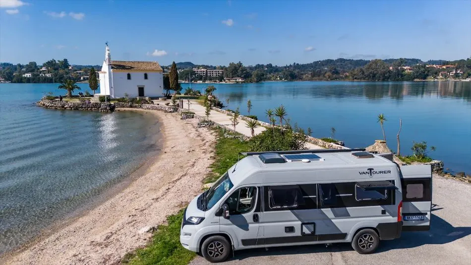 Deniz kenarındaki beyaz badanalı bir Yunan şapelinin yanındaki kumsalda park etmiş kompakt karavan