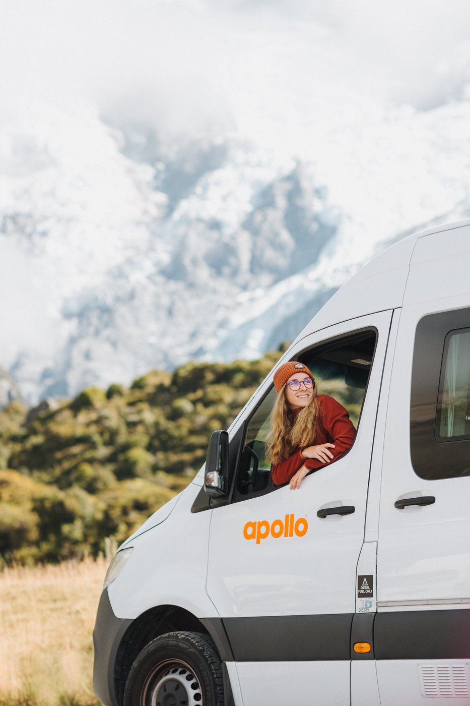 En leende resenär som lutar sig ut genom fönstret på en Apollo-husbil, med Aoraki/Mount Cook och Södra Alperna i bakgrunden
