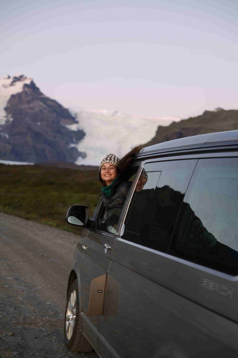 Two smiling travellers leaning out of a grey campervan window, with an Icelandic glacier and mountains behind